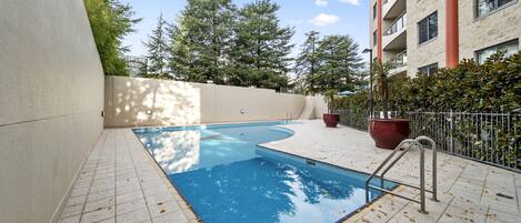 A swimming pool with a tiled deck, surrounded by a low wall and trees.