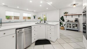 Una cucina moderna con mobili bianchi, lavastoviglie, lavello e forno integrato. Sono presenti piante in vaso e uno scaffale con bicchieri.