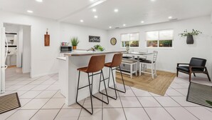 Una cucina moderna con mobili bianchi, un'isola centrale con sgabelli, un'area pranzo con tavolo e sedie, e vista sull'esterno attraverso una finestra.