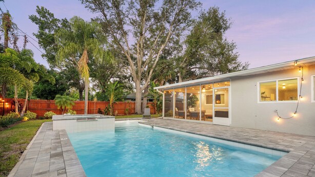 Una piscina con bordo in pietra, recinzione in legno e una casa con ampie finestre e luci decorative.