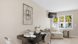 Un comedor moderno con una mesa de madera oscura, sillas blancas y una decoración minimalista. Un televisor de pantalla plana está montado en la pared, y una ventana ofrece vistas a la calle.