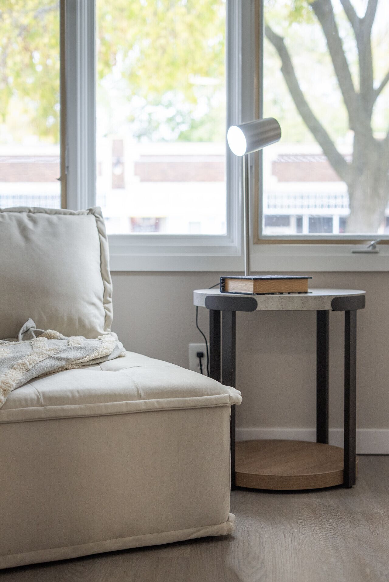 A room with a sofa, a table with a lamp, and a window with a view of trees and buildings.