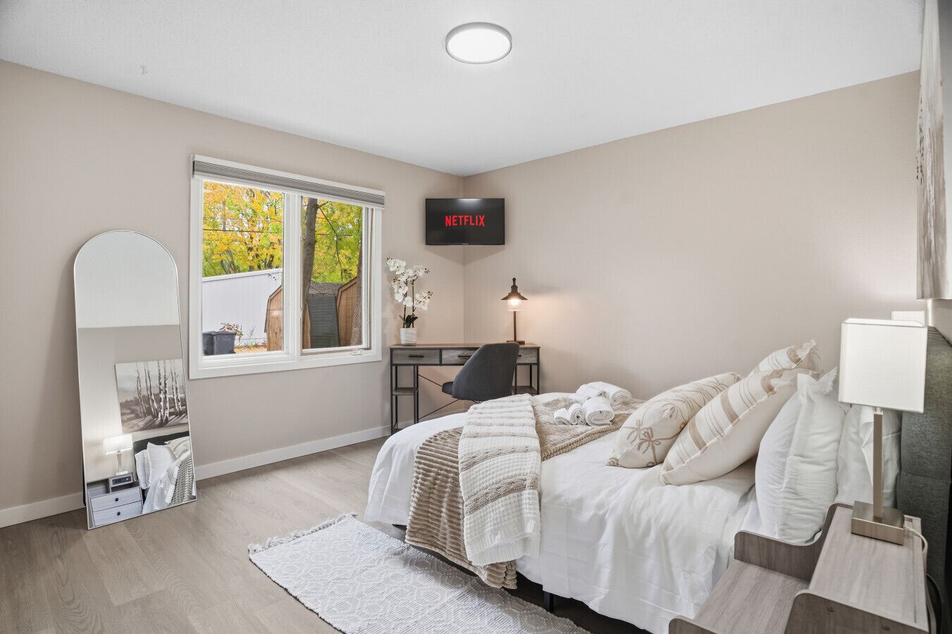 Un dormitorio con cama, escritorio, silla, lámpara, espejo y ventana con vista a árboles.