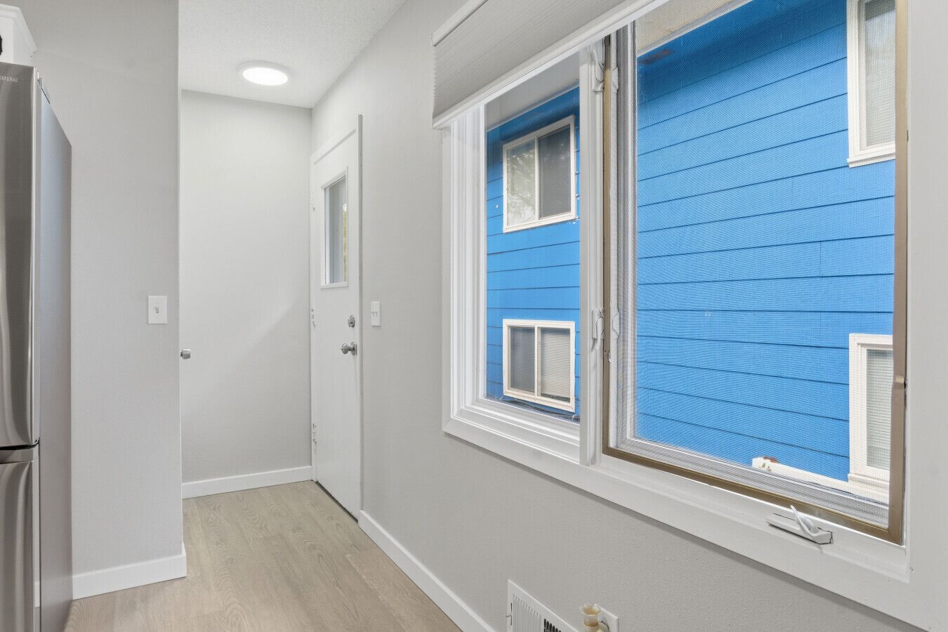 A narrow hallway with a refrigerator on the left, a door at the end, and a window with blue siding on the right.