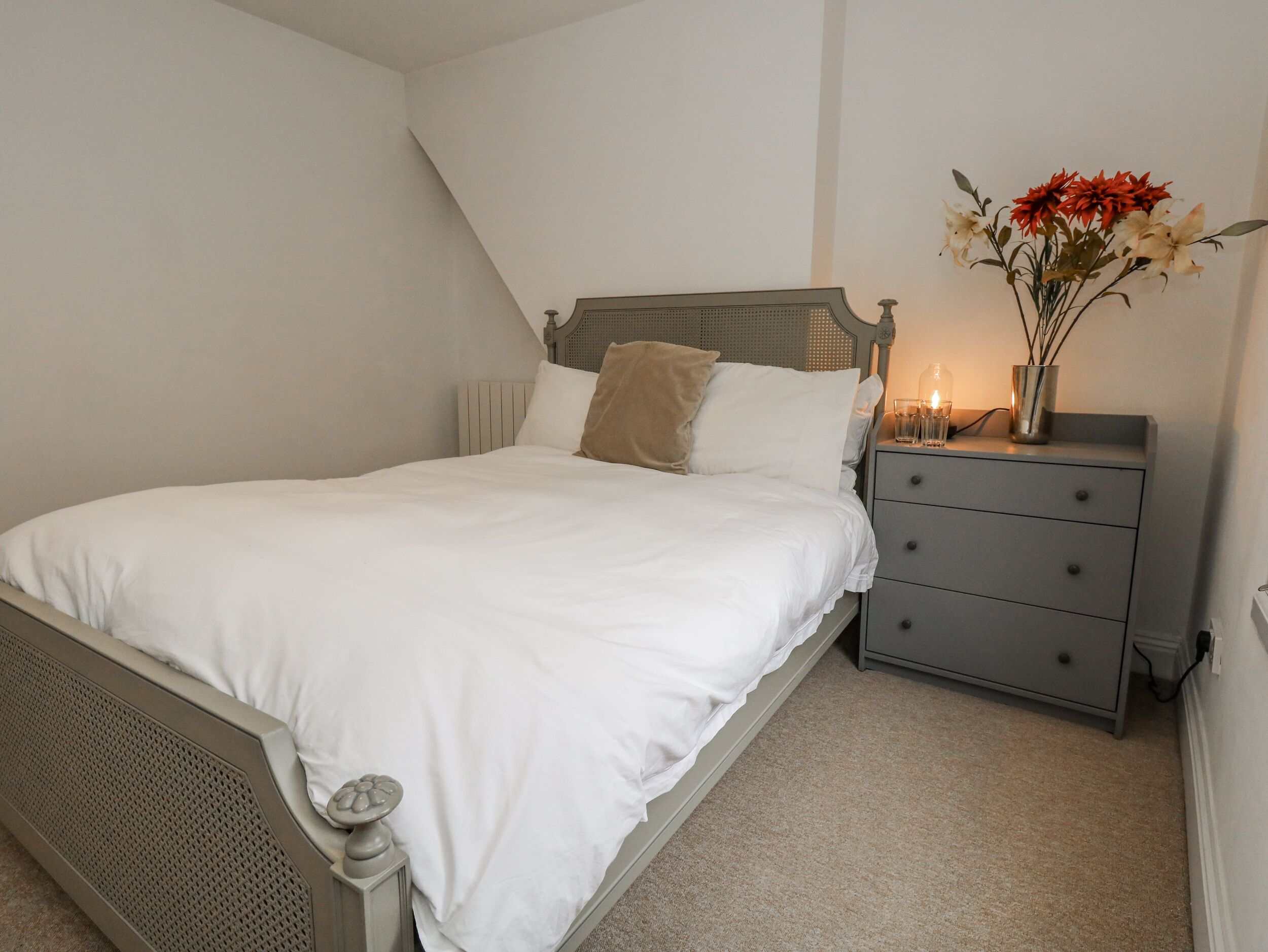 A bedroom with a bed, a nightstand, a vase with flowers, and a wall-mounted light.
