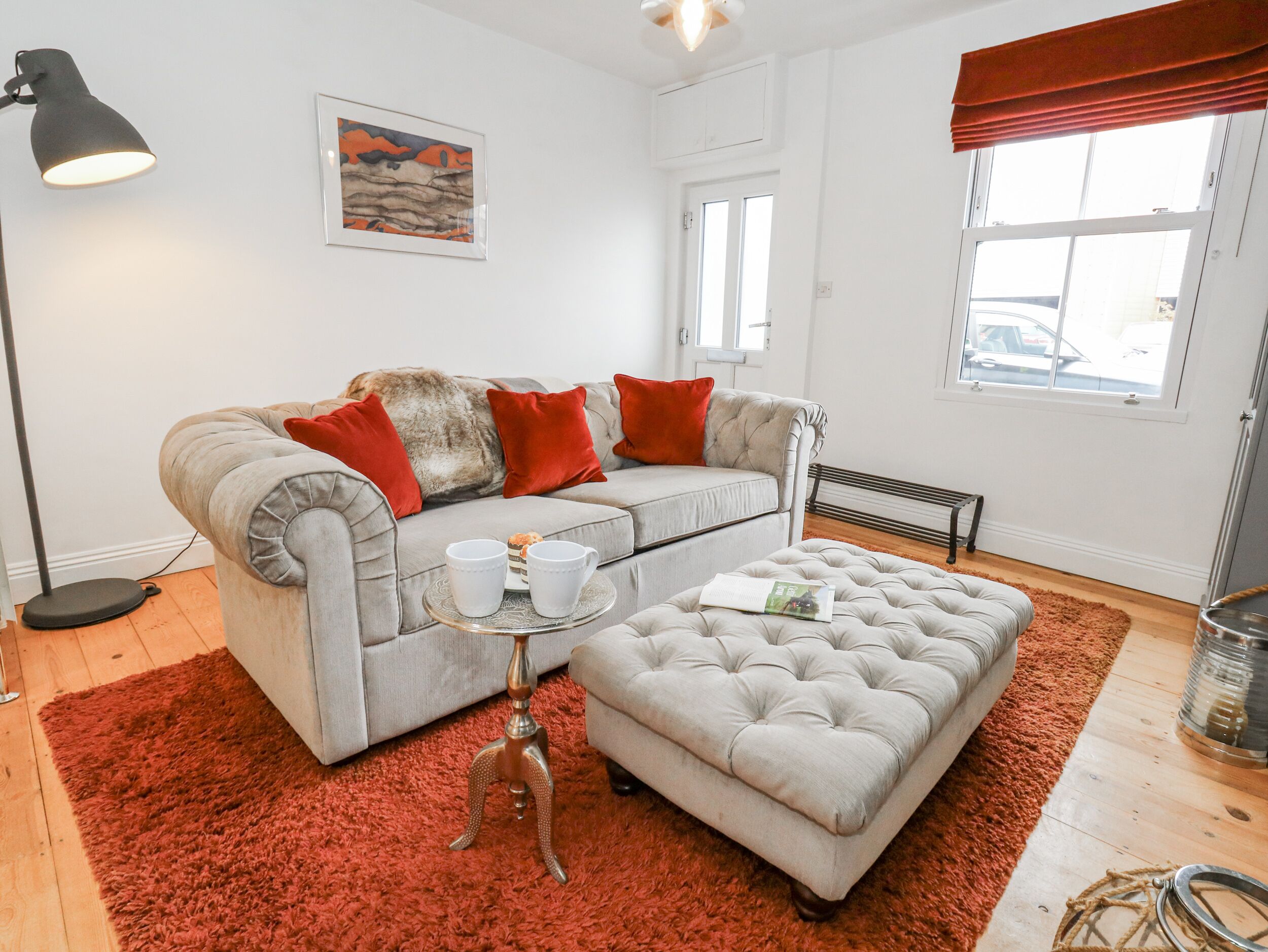 A living room with a grey sofa, red cushions, a small side table with cups and a tray, a red rug, and a window with a view of the outside.