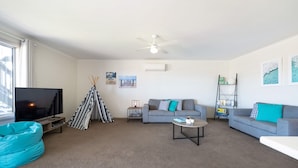 A modern living room with a flat-screen TV, a grey sofa, a blue bean bag, a small coffee table, and a ceiling fan. - Spacious 3-bed Ground Level Beachside Stay (Middleton)