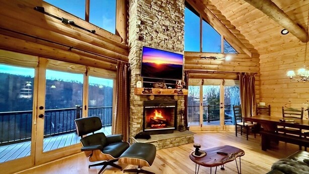 Ein Holzblockhaus-Interieur mit Kamin aus Stein, einem an der Wand befestigten Fernseher und einem Balkon mit Blick auf die umliegende Landschaft.
