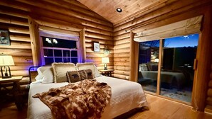 Ein Schlafzimmer in einer Blockhütte mit einem großen Bett, zwei Nachttischlampen, einem Fenster mit Aussicht und einer Glastür zum Außenbereich.
