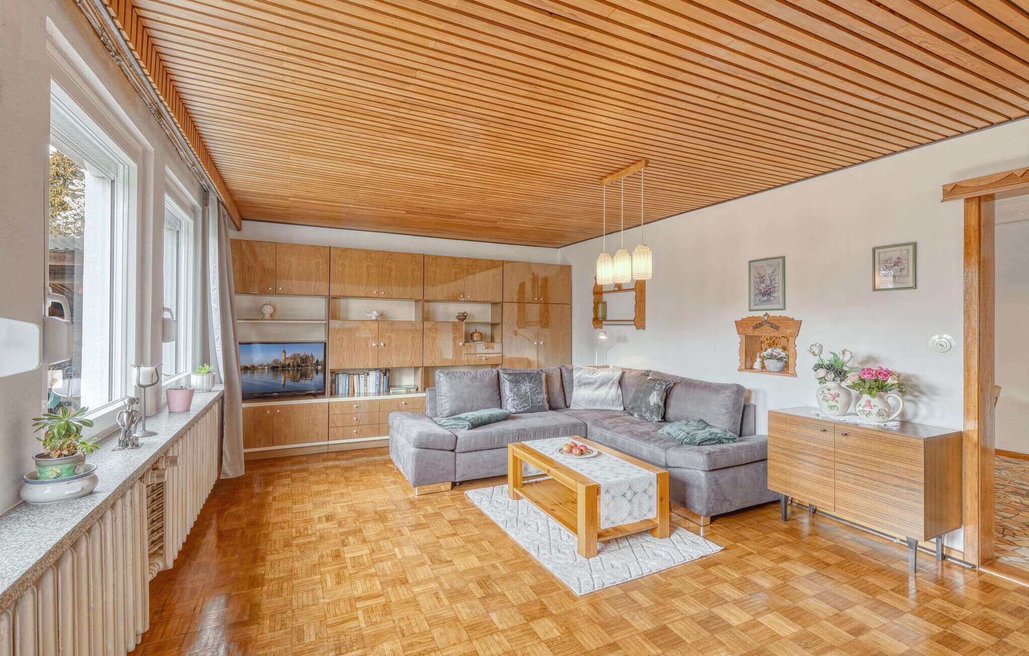 A spacious living area with a wooden ceiling, built-in wooden cabinets, a grey sofa, a wooden coffee table, and a view of a cityscape through large windows.