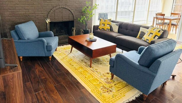 Ein Wohnzimmer mit Kamin aus Backstein, blauen Sofas, einem Holztisch und einem gelb gemusterten Teppich.