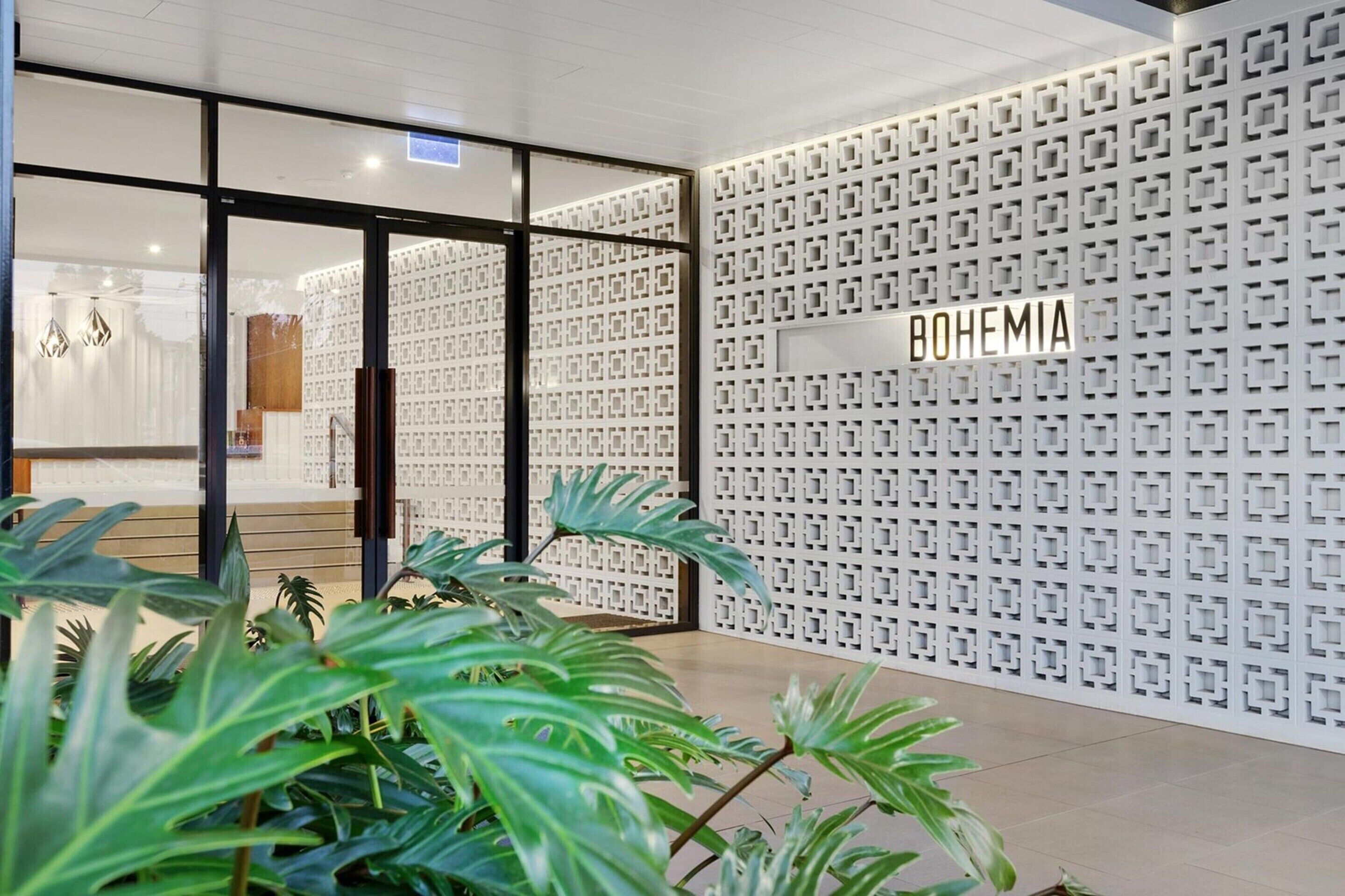 A modern interior with a glass door entrance, a patterned wall with the word "BOHEMIA" on it, and indoor plants.