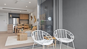 Een moderne woonkamer met twee witte frame stoelen, een ronde salontafel en een bank. Een glazen deur leidt naar een keuken met houten kasten en een eethoek.