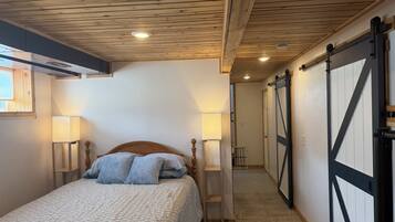 A bedroom with a wooden ceiling, a bed with a headboard, two bedside lamps, and a sliding barn-style door.