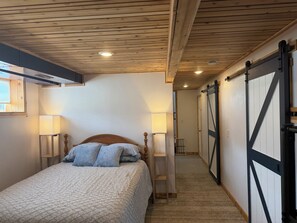 A bedroom with a wooden ceiling, a bed with a headboard, two bedside lamps, and a sliding barn-style door.