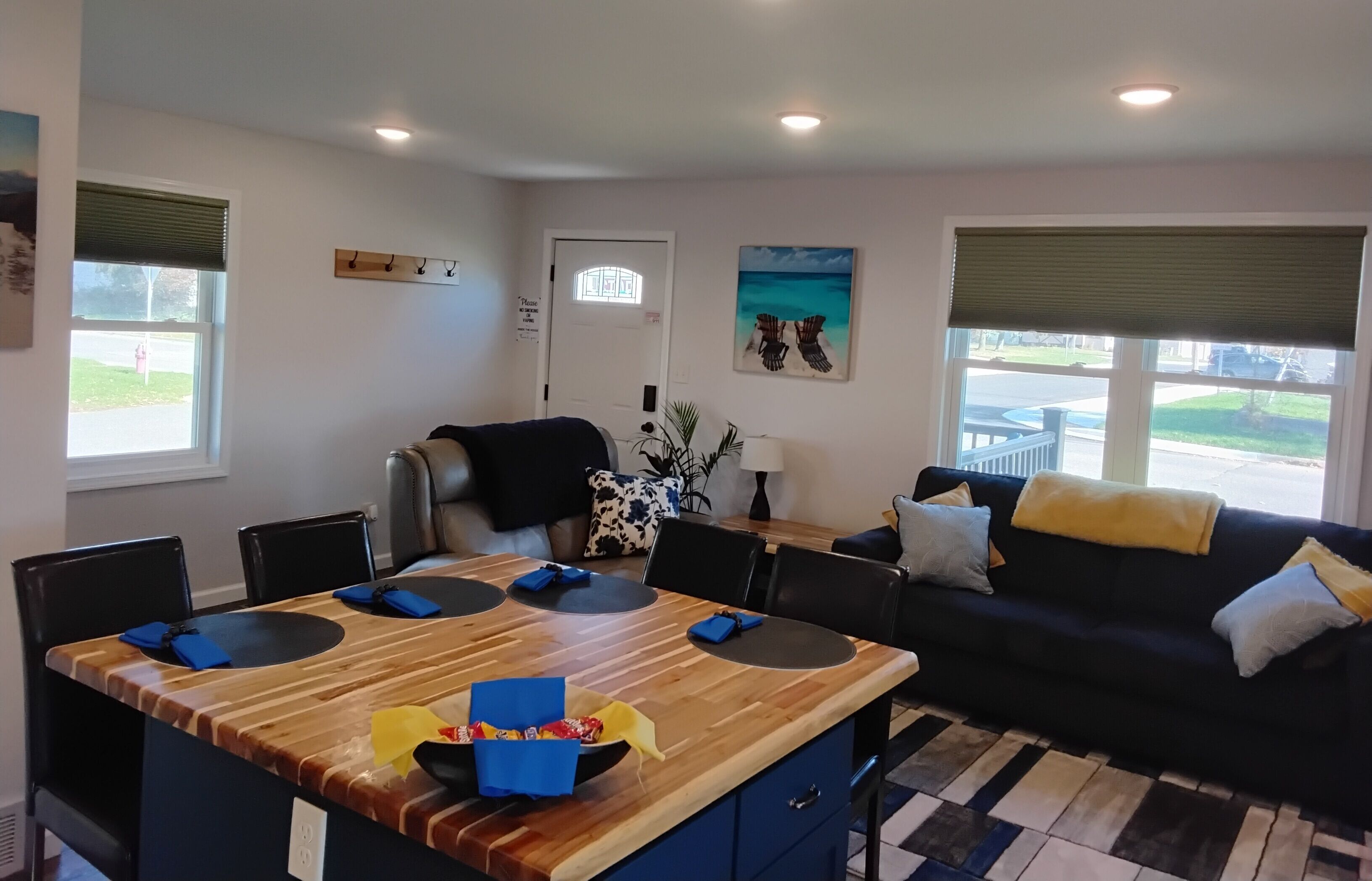 Een moderne woonkamer met een houten eettafel, zwarte stoelen, een zwarte bank met gele en witte kussens, en uitzicht op de straat door grote ramen.