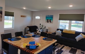 Een moderne woonkamer met een houten eettafel, zwarte stoelen, een zwarte bank met gele en witte kussens, en uitzicht op de straat door grote ramen.