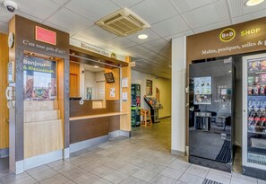 Zona de recepción de un hotel con mostrador de madera, una mesa de recepción y una tienda con aperitivos y bebidas.