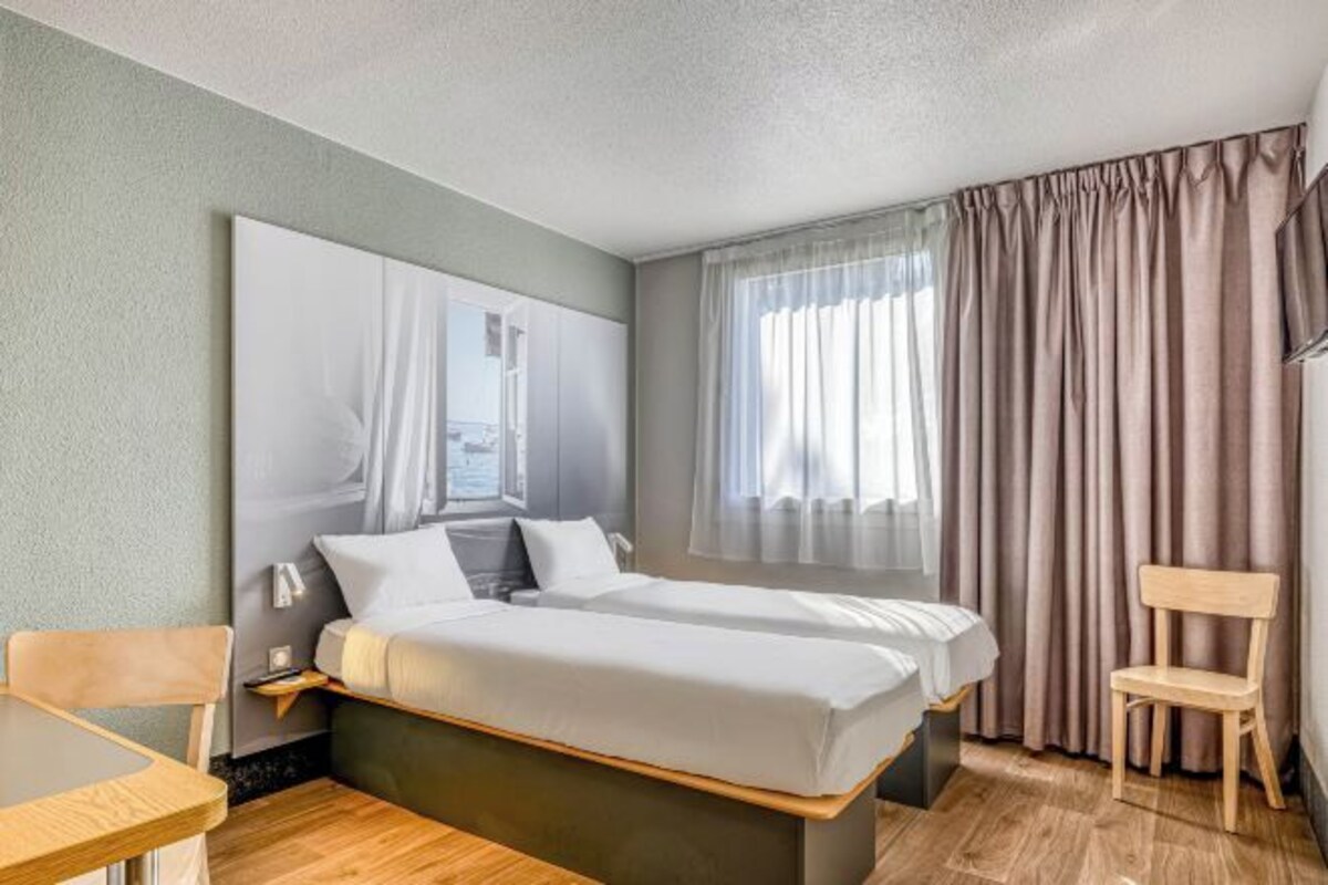 Una habitación de hotel con cama, escritorio, silla y un gran ventanal con cortinas.