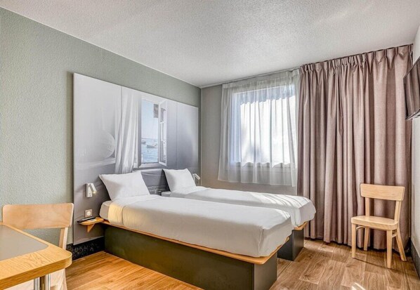 Quarto de hotel com uma cama, mesa de cabeceira, cadeira, janela com cortinas e uma obra de arte na parede.