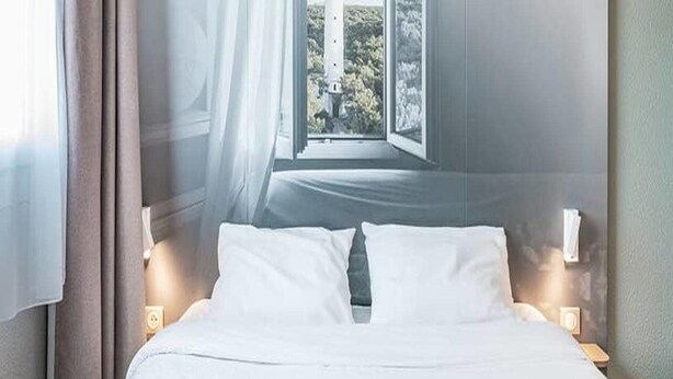 A hotel room with a bed, a window with a view of greenery, and a nightstand with a lamp. - B&B Hotel Bordeaux Sud Mios (Mios)