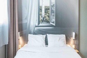 Quarto de hotel com cama, janela com vista para vegetação e mesa de cabeceira com luminária.