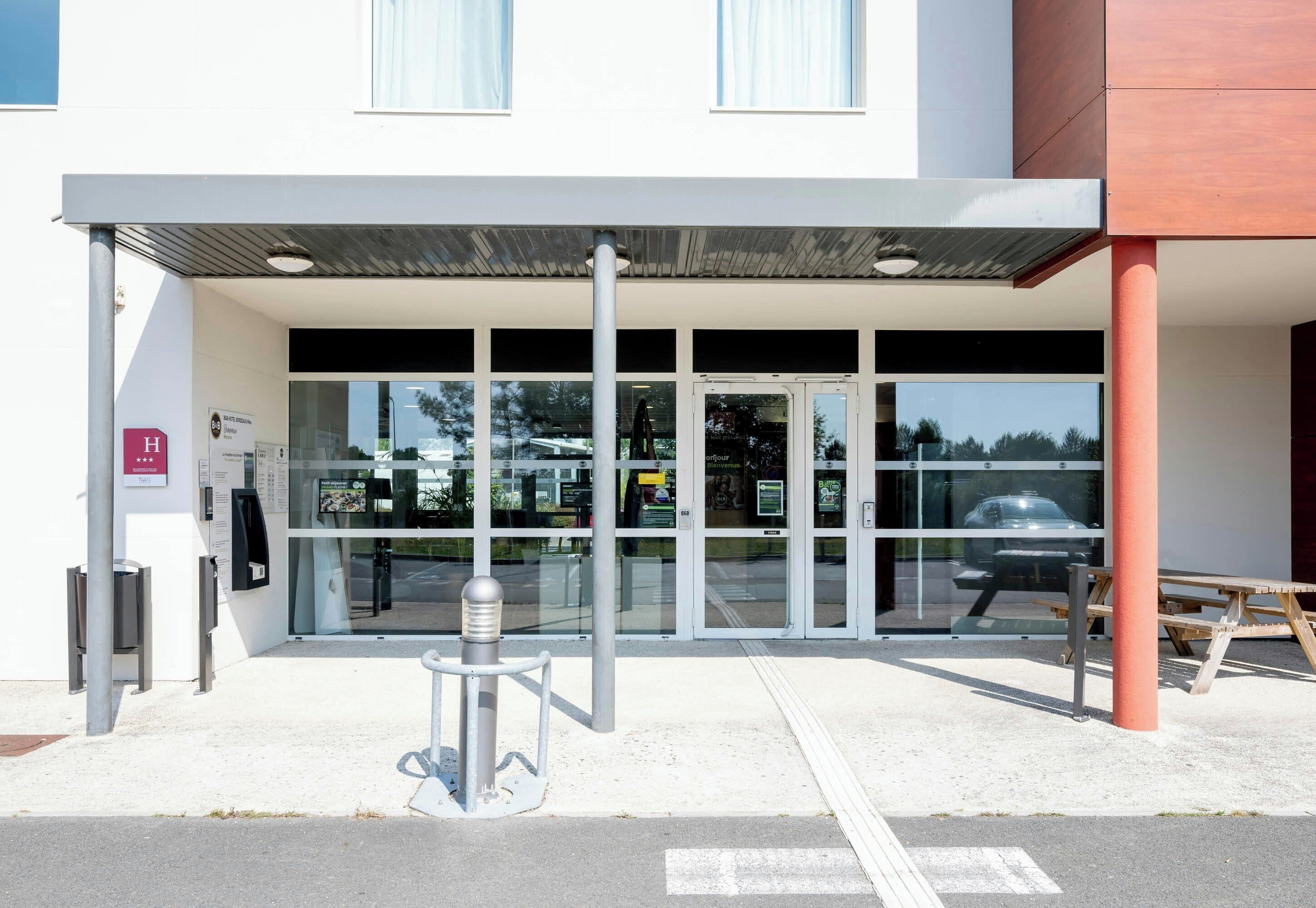 Entrada de um edifício moderno com galeria coberta, portas de vidro e um carro visível ao fundo.