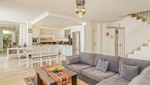 Un vaste salon avec un canapé gris, une table à manger en bois et des chaises. On aperçoit une cuisine à l’arrière-plan, avec des meubles blancs et un réfrigérateur en acier inoxydable.