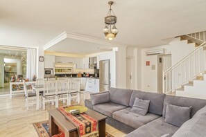 A spacious living area with a grey sofa, a wooden coffee table, and a dining table with chairs. A kitchen is visible in the background, and a staircase leads to another level.