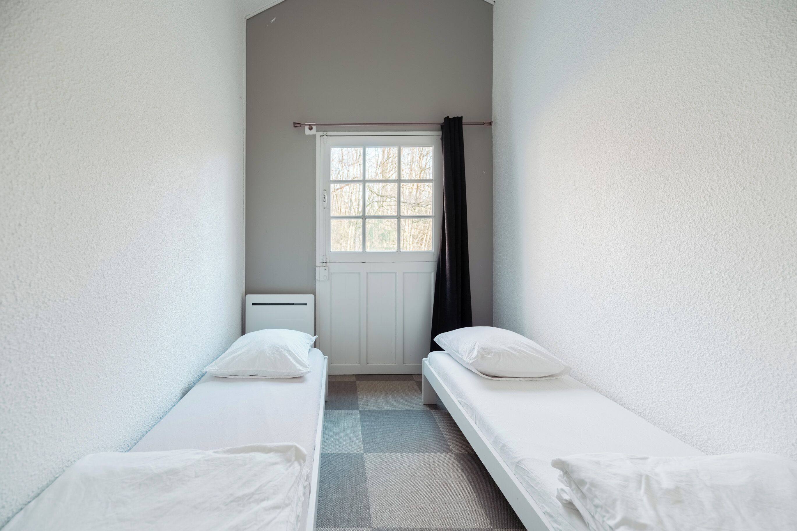 Ein kleines, sauberes Zimmer mit zwei Einzelbetten, einem Fenster mit Vorhang und einer Heizung.