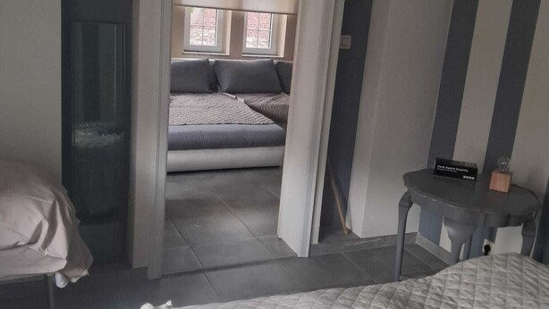 Ein Schlafzimmer mit Bett, Nachttisch, Stuhl und Blick auf einen anderen Raum mit Sofa und Fenstern.