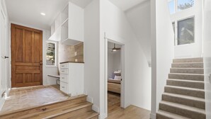 Ein modernes Interieur mit einer Holztreppe, einer Küchenzeile mit weißen Schränken und einem Schlafzimmer mit Bett und Fenster.