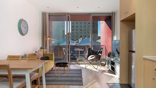 A modern living room with a dining area, a sofa, a chair, and a table. There is a large window with a view of buildings outside. - Chic Apartment in Heart of Adelaide (Adelaide)
