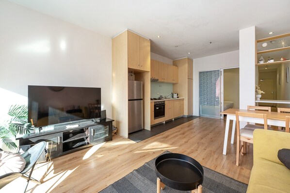 A modern living room with a flat-screen TV, a dining area with a table and chairs, a kitchen with appliances, and a large window. - Chic Apartment in Heart of Adelaide (Adelaide)