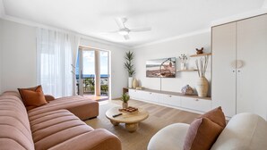 Een moderne woonkamer met een bank, fauteuil en salontafel, een plafondventilator en uitzicht op de omgeving.