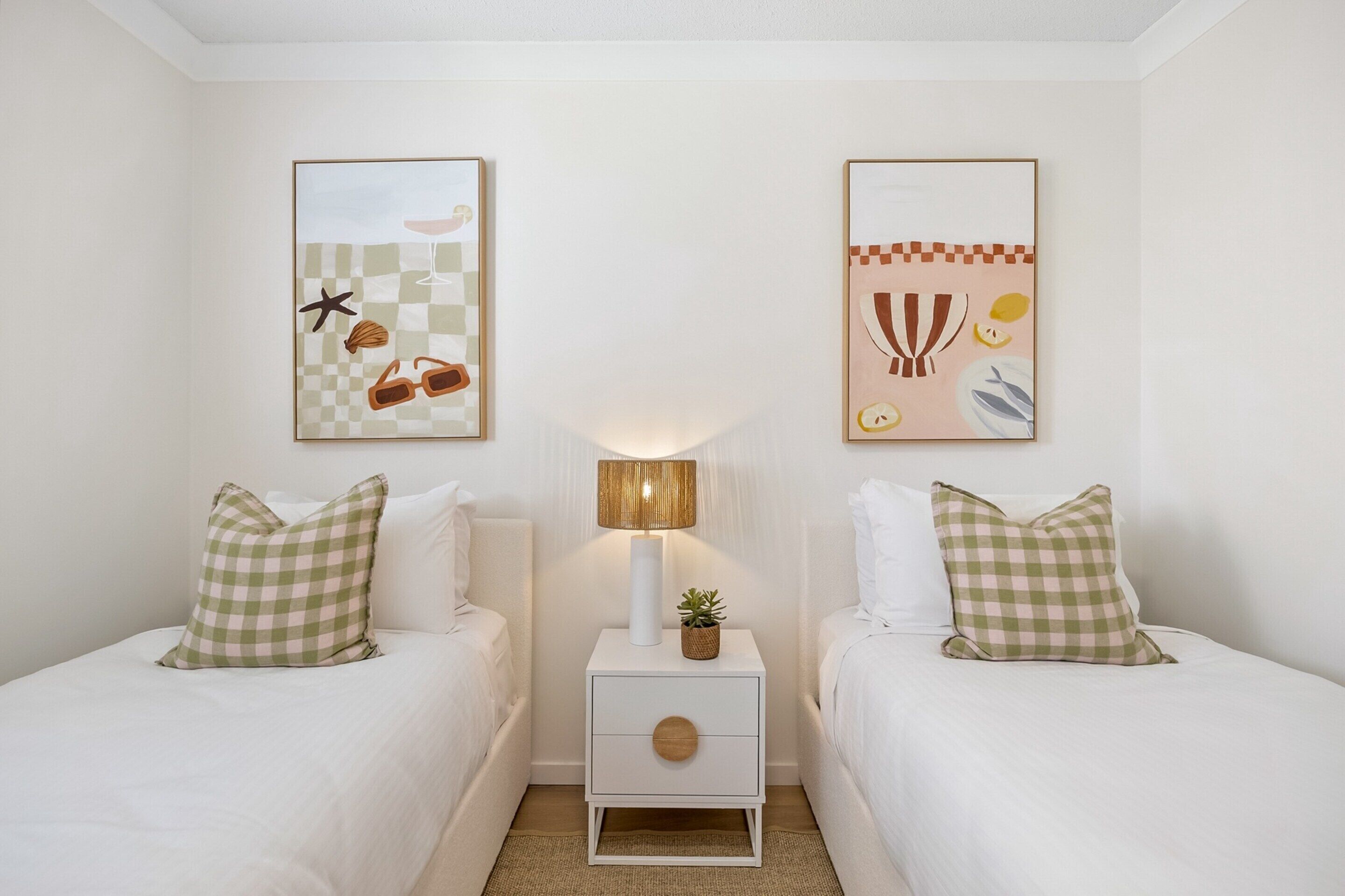 A hotel room with two beds, a nightstand with a lamp, and two wall-mounted artworks.