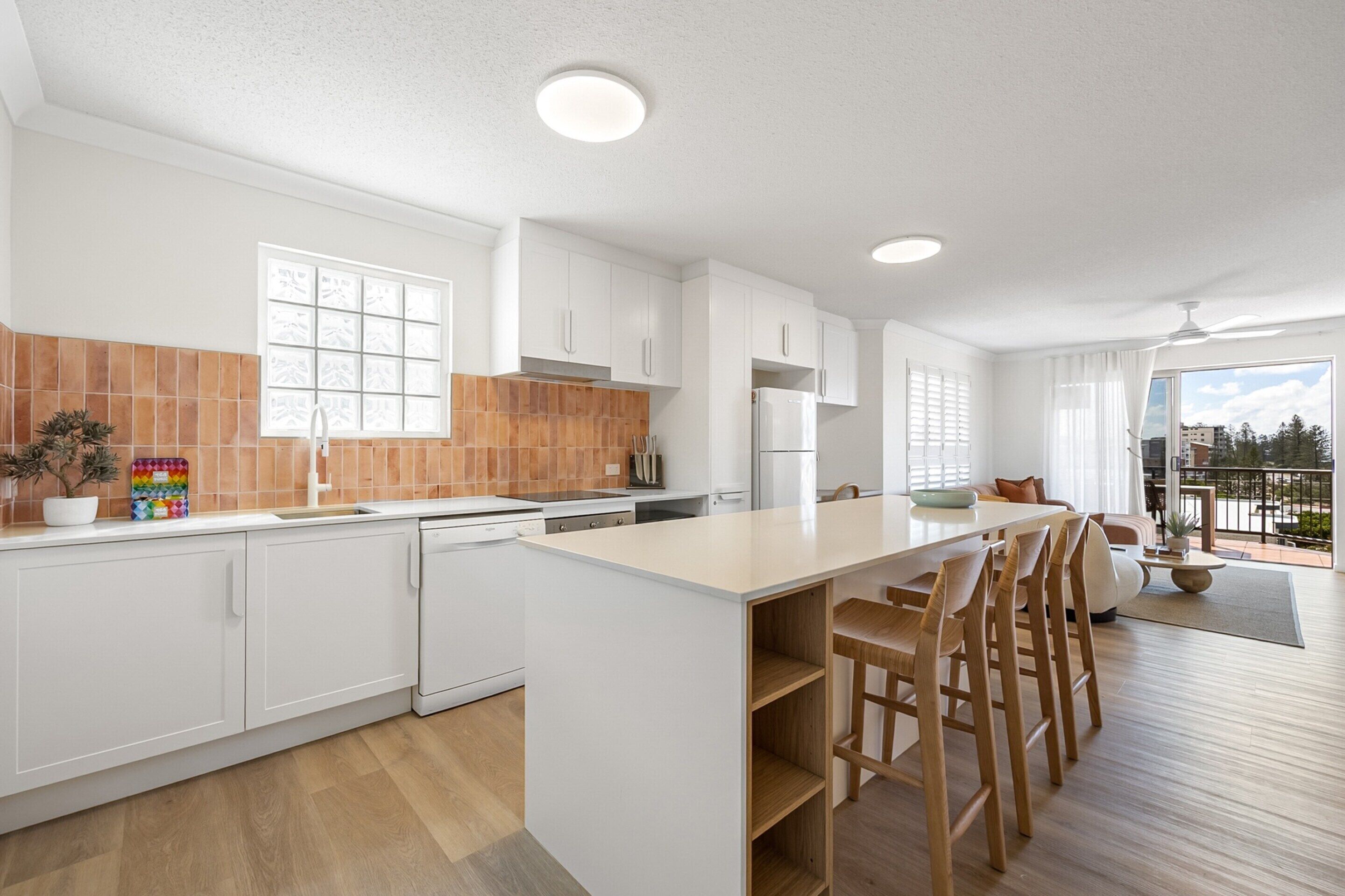 A modern kitchen with white cabinets, a wooden island with bar stools, a large window, and a view of the outdoors.