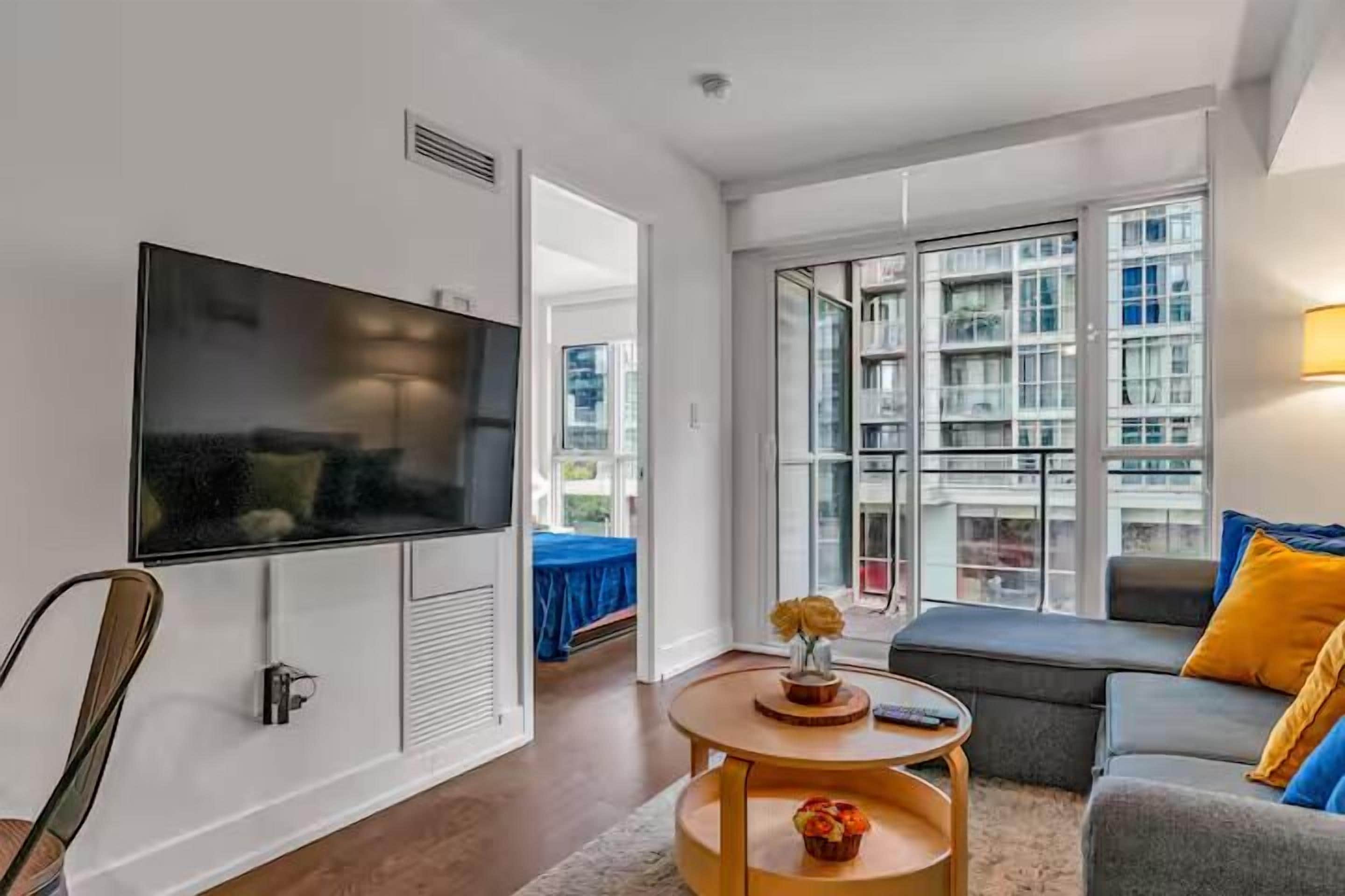 A modern living room with a flat-screen TV, a sofa, a round coffee table, and a large window offering a view of the city.