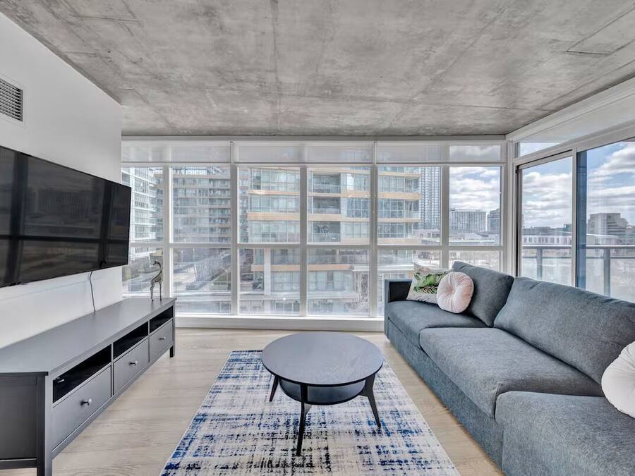 Een moderne woonkamer met een grote televisie, een grijze bank en uitzicht op stadsgebouwen door grote ramen.