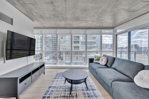 Un salon moderne avec un grand téléviseur, un canapé gris et une vue sur les immeubles de la ville, visible à travers de grandes fenêtres.