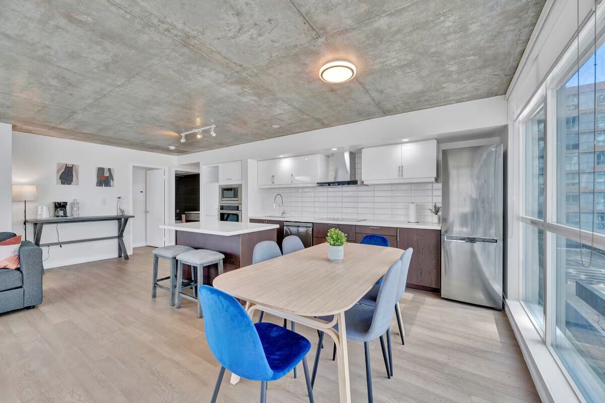 A modern kitchen with white cabinets, a stainless steel refrigerator, and a dining area with a wooden table and blue chairs.
