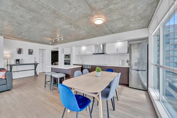 A modern kitchen with white cabinets, a stainless steel refrigerator, and a dining area with a wooden table and blue chairs.