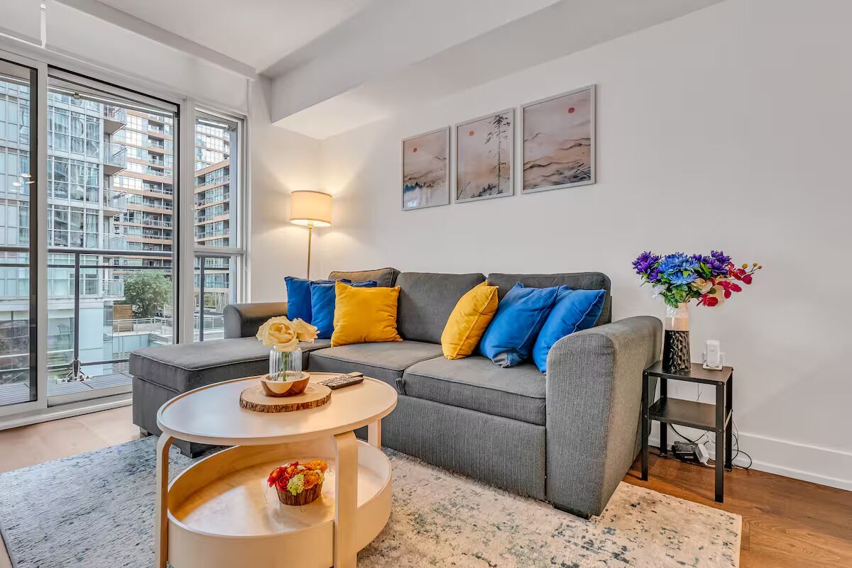 A modern living room with a grey sofa, yellow and blue cushions, a round coffee table, and a side table with a vase of flowers.