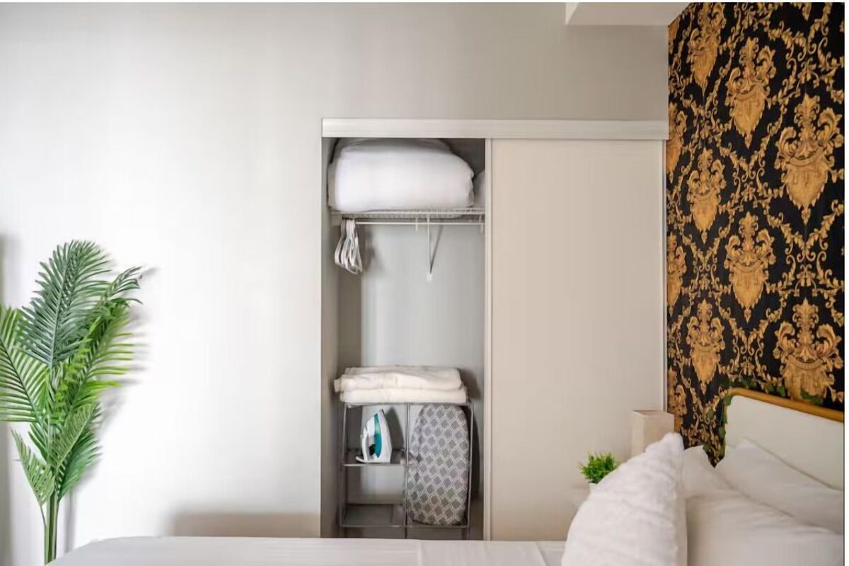 A hotel room with a bed, a wardrobe with folded clothes, an ironing board, and a potted plant.