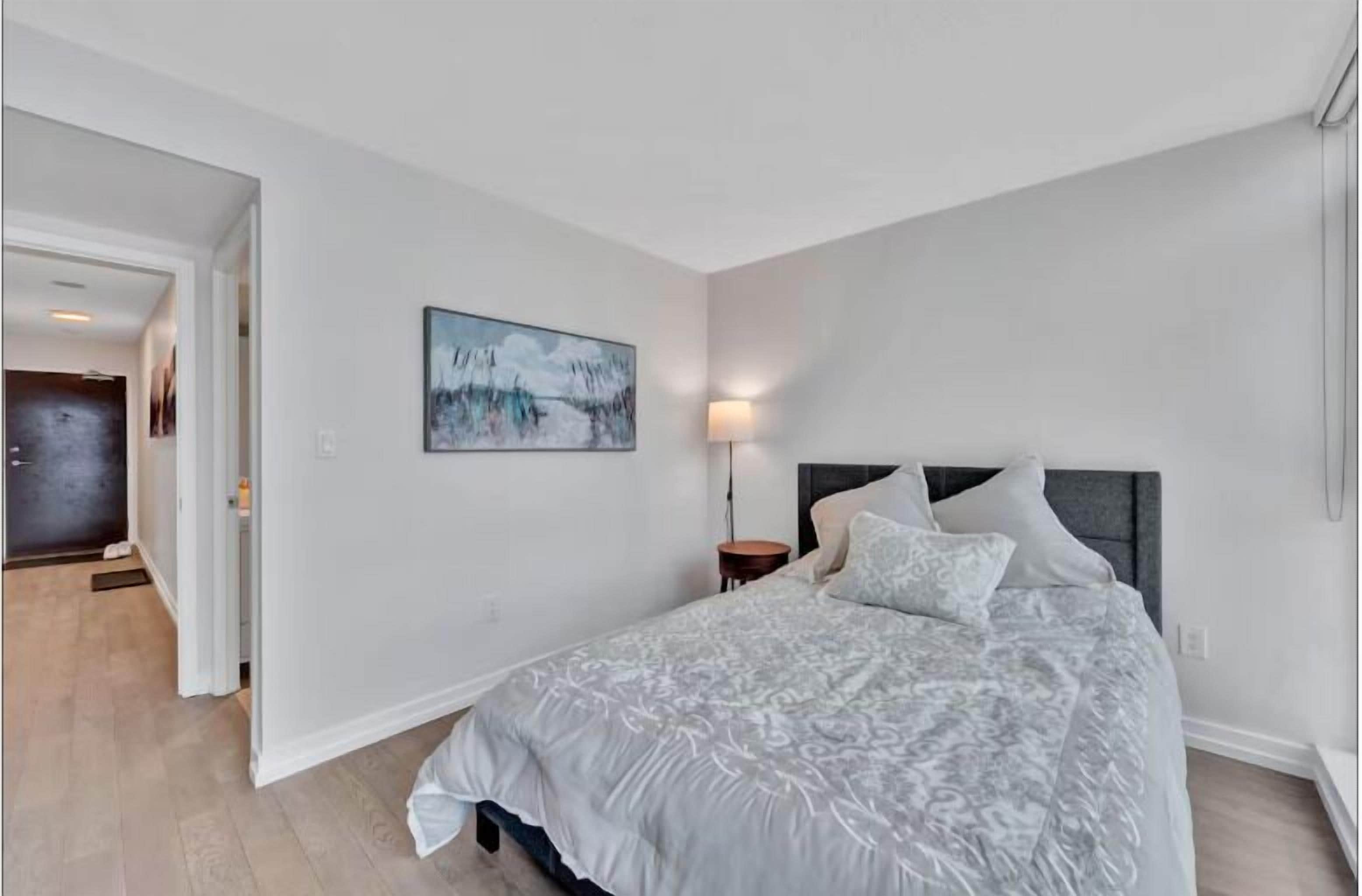 A bedroom with a large bed, a nightstand, a wall-mounted lamp, and a painting.