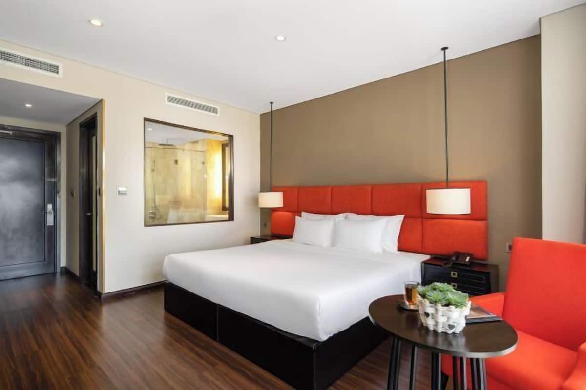 Ein Hotelzimmer mit einem großen Bett, einem rot gepolsterten Kopfteil, einem kleinen Tisch mit einer Pflanze und einem Sessel.