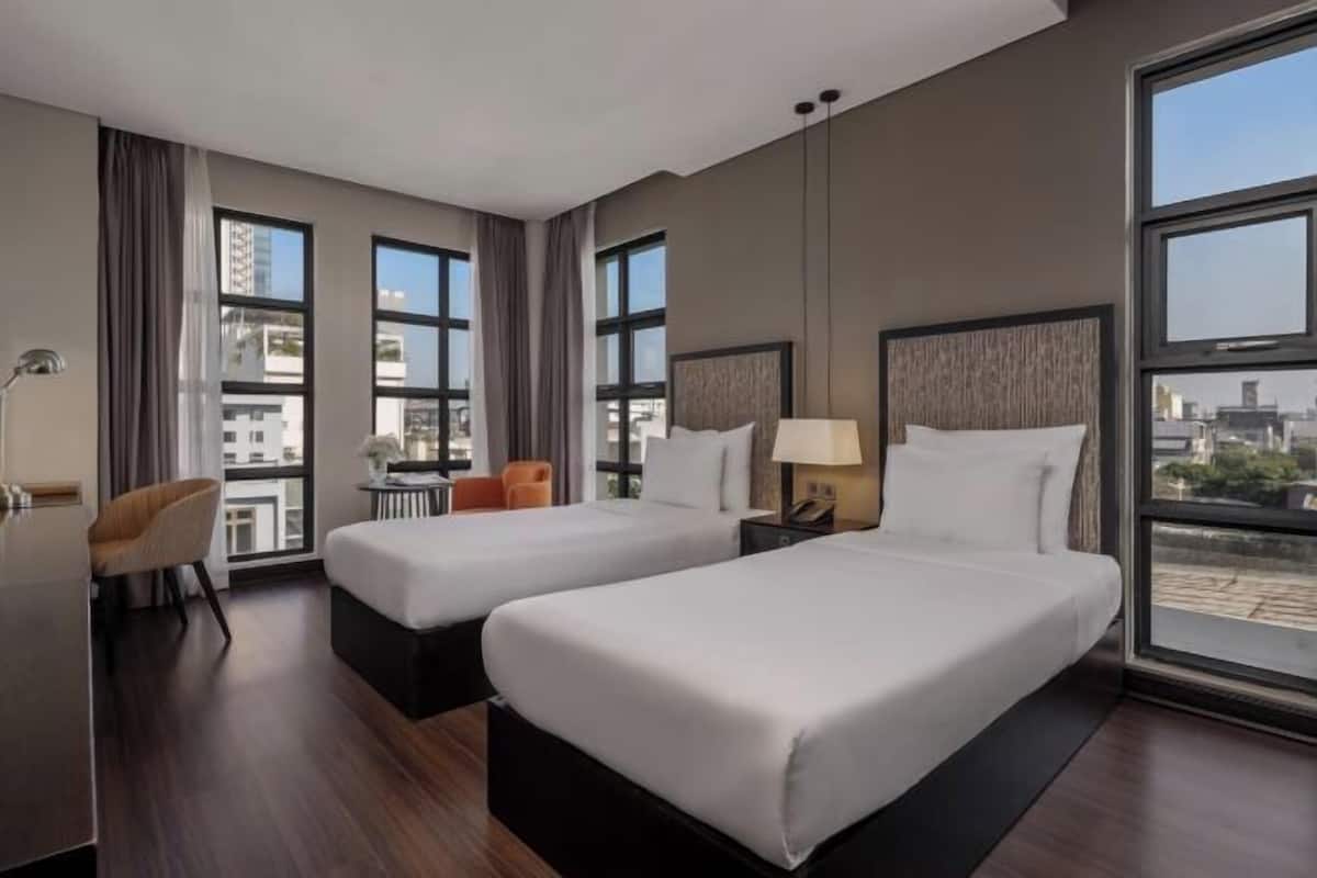 Ein Hotelzimmer mit zwei Betten, einem Schreibtisch, einem Stuhl und großen Fenstern mit Blick auf die Stadt.