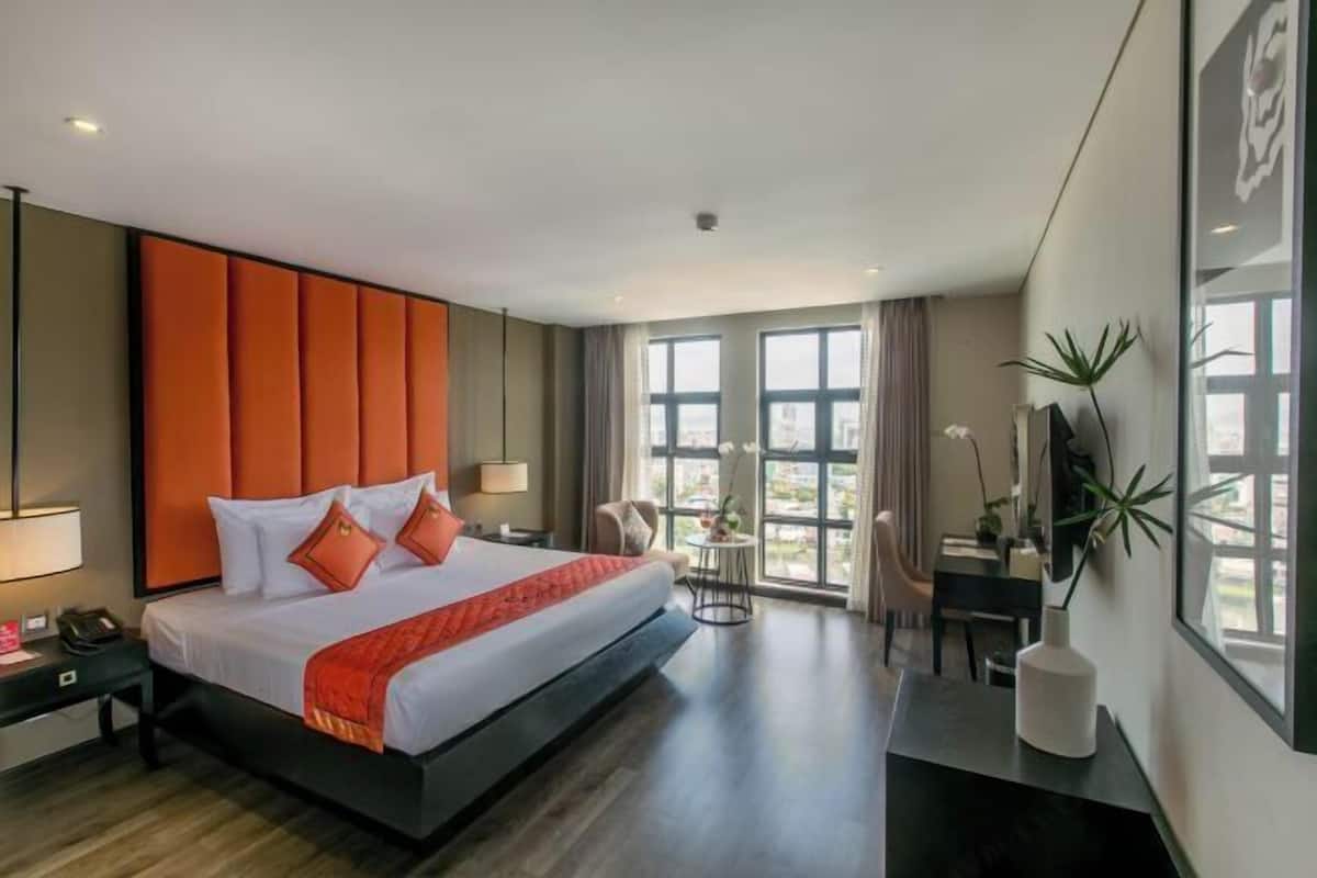 Ein modernes Hotelzimmer mit einem großen Bett, einem Schreibtisch, einem Sessel und Blick auf die Stadt durch große Fenster.