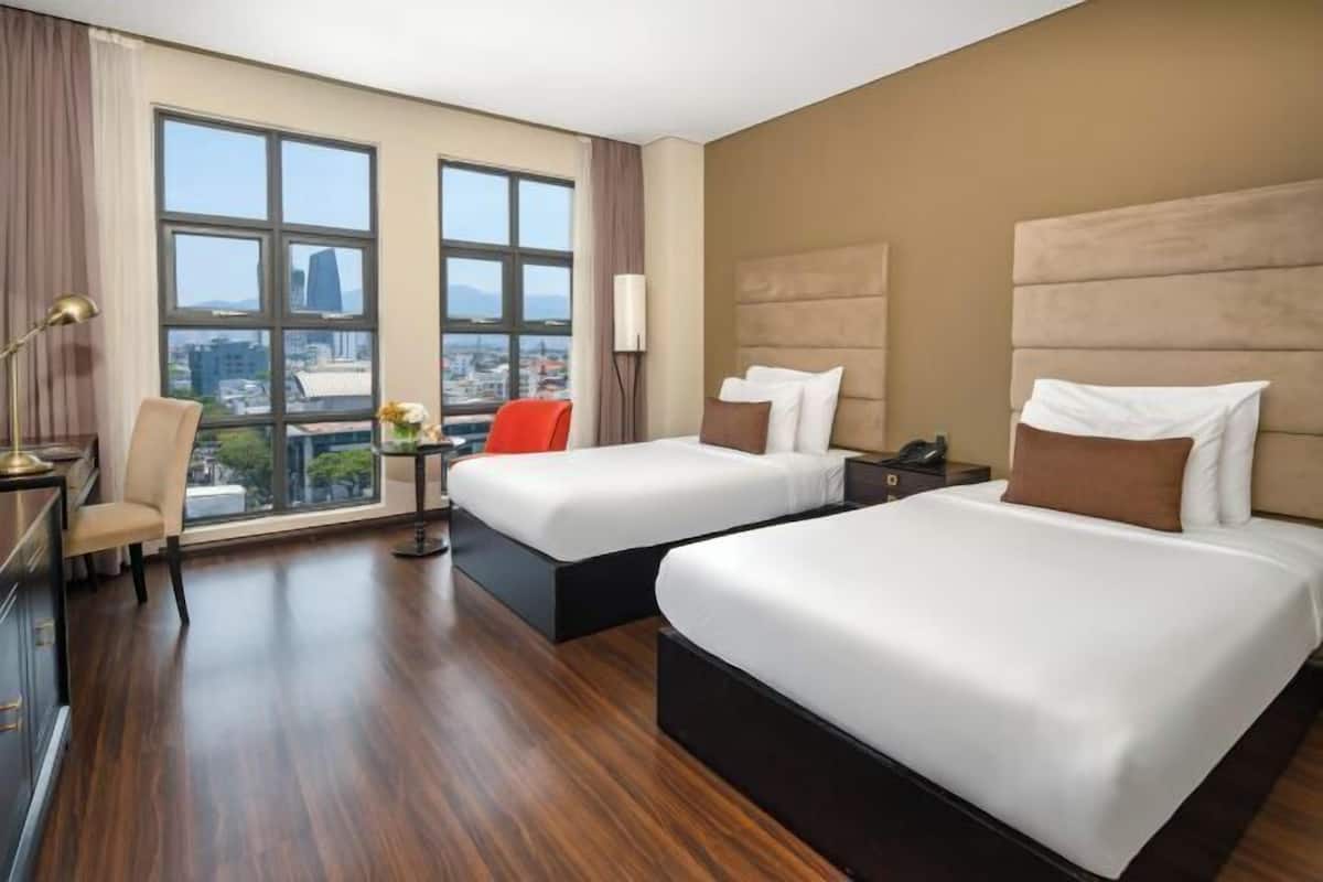 Ein Hotelzimmer mit zwei Betten, einem Schreibtisch, einem Stuhl und einem großen Fenster mit Blick auf die Stadt.