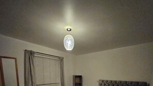 Un dormitorio con cama, cómoda, radiador, ventana con cortinas e iluminación en el techo.